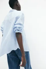 Camisa celeste de lino liviano, con cuello, botones al frente, canesú de doble capa en la espalda, hombros caídos, mangas largas con puños abotonados, bolsillo superior de parche y bajo levemente redondeado.