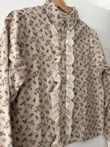 Camisa color beige con estampado floral, cuello camisero, mangas largas abullonadas y cierre frontal con botones. Presenta detalle de puntilla en el frente.