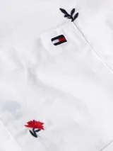 Camisa blanca de manga larga con cuello clásico y bolsillo frontal. Presenta un estampado repetitivo de pequeñas flores bordadas en tonos rojo, azul y negro. El interior del cuello tiene un detalle de rayas azules y blancas.