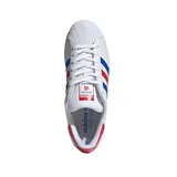Championes Adidas Superstar blancos con detalles en azul y rojo. Presentan las tres tiras características de Adidas en los laterales en azul y rojo, y refuerzo de talón en rojo con la palabra "STAR" en blanco. La puntera es de goma con textura.