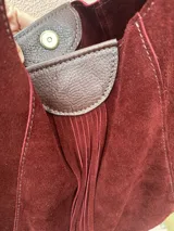 Cartera tipo hobo de gamuza color borgoña con flecos y asa corta de cuero marrón.