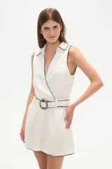 Mono corto color blanco, sin mangas, con cuello camisero, corte cruzado y cinturón con hebilla circular.