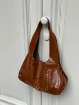 Cartera tipo hobo de cuero color marrón, con bolsillo externo y tachas plateadas.