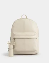 Mochila de cuerina color crudo con bolsillo frontal con cierre y charm decorativo con cierre. Cuenta con correas regulables y manija superior.