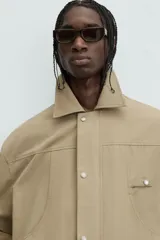 Cazadora color arena de corte relaxed fit, confeccionada en tejido de algodón. Cuenta con cuello solapa, manga larga con bolsillo multifunción, bolsillos frontales de plastrón, bolsillo interior, acabados con elástico y bajo ajustable con cordón. Cierre frontal con cremallera oculta por solapa con botones a presión.