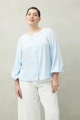 Camisa celeste de corte holgado con botones frontales, canesú con tablas y mangas 3/4 amplias con puño elastizado.