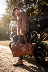 Bolso de cuero marrón hecho a mano con diseño tipo duffle. Cuenta con asas de mano reforzadas, correa larga regulable y extraíble, compartimento principal con cierre y bolsillo exterior frontal.