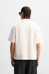 Camisa color blanco con textura de rayas verticales, corte relaxed fit, cuello bowling, manga corta, bolsillo de parche en el pecho y cierre frontal con botones.