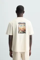 Camiseta color crudo de corte relaxed fit, con cuello redondo y mangas cortas. Presenta un pequeño bordado de flores en el pecho y un estampado fotográfico en la espalda.