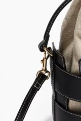 Cartera bombonera de cuero sintético color beige con apliques de tiras de cuero sintético negro y costuras blancas. Tiene doble asa corta y correa larga extraíble. El interior es de lona con cordón ajustable.