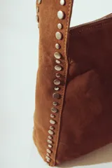 Bolso de hombro tipo hobo, color camel, confeccionado en gamuza o material similar con acabado afelpado. Presenta detalles de tachas metálicas plateadas a lo largo de los bordes laterales y en la correa de hombro.