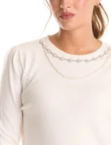 Buzo tejido en hilado fino, color blanco, con escote a la base y un collar decorativo de perlas y strass. Tiene mangas largas con un leve frunce en los hombros.