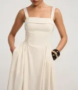 Vestido midi de corte evasé, color crema, con escote cuadrado y breteles anchos. Presenta un estampado grande de hojas y follaje en tonos oscuros en el ruedo inferior.