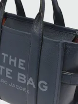 Bolso tote pequeño de cuero texturizado color gris con doble asa corta superior y correa larga extraíble y ajustable para llevar al hombro. Estampado frontal con el texto "The Tote Bag" y el nombre de la marca.