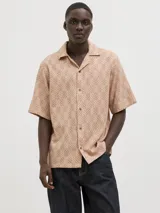 Camisa de manga corta Jack & Jones con cuello resort y cierre de botones. Diseño con estampado de espiga en tonos marrones y blancos. Confeccionada en tejido dobby 100% algodón.