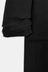 Blazer negro de corte recto, con cuello solapa con muescas, hombreras y un solo botón dorado frontal. Las mangas son de largo tres cuartos con detalle de drapeado o fruncido.