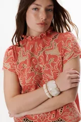 Blusa color coral o terracota con bordado calado floral en tono beige claro. Presenta cuello alto con volado y mangas cortas abullonadas.