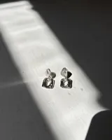 Par de aros colgantes de plata 950, con diseño orgánico y textura martillada. Cada aro consta de dos piezas unidas: una superior redondeada y una inferior cuadrada con un orificio central.