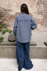 Camisa de rayas verticales azules y blancas, con cuello camisero, manga larga y puños abotonados. Presenta un bolsillo en el pecho con ribete rojo y un detalle de ribete rojo en el cuello y la tapeta de botones.