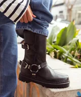 Bota de caña media color marrón oscuro, realizada en cuero encerado. Con hebillas en la caña y herrajes de níquel. El fondo está realizado artesanalmente, es de suela, y es negro. El interior está forrado en forrería vacuna.