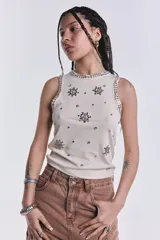 Musculosa color crudo de punto canalé, con cuello redondo y sisas ribeteadas con strass. Presenta apliques de strass con forma de flor y tachas plateadas dispersas por toda la prenda.