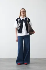 Chaleco de punto color blanco con bordado aplicado de cordón marrón en diseño floral. Presenta cierre frontal de botones al tono, cuello redondo y terminaciones acanaladas en sisas y ruedo.