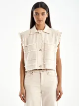 Chaqueta vaquera color crema, sin mangas, con cuello camisero, cierre frontal con botones metálicos, dos bolsillos frontales con botón y detalle de borde deshilachado.