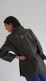 Blazer de cuero color gris oscuro, con aberturas laterales, aplique tipo pin desplegable bañado en bronce y botones de nácar.
