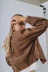 Campera bomber color beige de cuero, con cuello redondo, cierre frontal con botones a presión, mangas largas con puños elásticos y detalles de tachas metálicas en hombros y espalda.