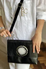 Cartera de mano tipo sobre, color beige claro, con solapa frontal decorada con una piedra de ágata azul engarzada en metal plateado. Posee correa larga ajustable de cuero beige.