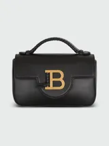 Bolso mini Balmain modelo B-Buzz, color negro, con asa superior rígida y correa de hombro extraíble. Presenta solapa con cierre de broche a presión y aplique metálico dorado con el logo de la marca.