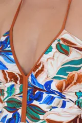 Traje de baño entero con estampado tropical de hojas en tonos azules, verdes y marrones sobre fondo blanco. Presenta escote en V profundo con ribete color marrón y frunces laterales para realzar la silueta. Se ata al cuello con tirantes finos color marrón.