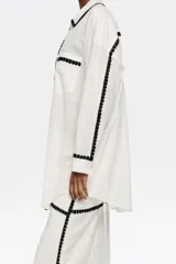 Camisa larga color blanco con borde de encaje negro, de corte recto, con cuello camisero, manga larga con puño y botón, y cierre frontal con botones.