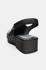 Zueco de piel negro con puntera redonda, plataforma de madera oscura y tachas doradas decorativas alrededor de la suela. Posee tira trasera ajustable con hebilla y cierre adhesivo.