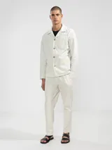 Pantalón de vestir color crema, confeccionado en tejido ligero de algodón con textura seersucker. Tiene corte recto, bolsillos laterales y traseros, cierre con botón y cremallera, y parche con logo de la marca.