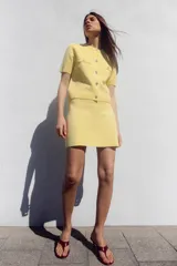 Chaqueta de punto color amarillo pálido, con cuello redondo y manga corta. Cierre frontal con botones dorados y bolsillos de parche en el pecho.