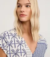 Enterito corto sin mangas de viscolino, con escote en V y estampado tipo patchwork en tonos blanco y azul marino, con motivos tropicales como palmeras y patrones geométricos. Incluye trabillas en la cintura y cinturón de cuerda trenzada.