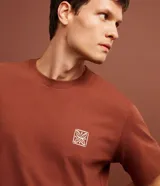 Remera color ladrillo de corte clásico, con cuello redondo y mangas cortas. Presenta un pequeño estampado cuadrado en el pecho.