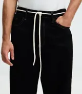 Pantalón masculino negro de corte baggy, confeccionado en sarga de algodón. Presenta detalles de costura, cierre tradicional de botón y cremallera, y un cordón blanco para ajustar la cintura.