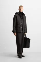 Parka negra de corte regular, confeccionada en tejido técnico acolchado. Tiene cuello alto con capucha ajustable, mangas largas con puños ajustables, bolsillos con solapa en la cadera y un bolsillo interior. El cierre frontal es con cremallera oculta por solapa con botones a presión.