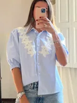 Camisa celeste con cuello clásico, mangas abullonadas 3/4 con puños elásticos y apliques de flores bordadas en el frente. Cierre frontal con botones. 