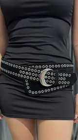 Cinturón ancho de cuero sintético negro con ojalillos metálicos plateados y hebilla plateada en forma de media luna.