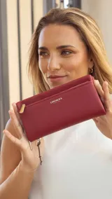 Billetera larga de mujer color rojo, con textura granulada, cierre de cremallera en el compartimento superior y un bolsillo exterior con cierre en el frente. Presenta un pequeño dije colgante de metal.