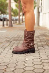 Bota corta de cuero color marrón, con taco cuadrado y punta fina. Presenta una correa con tachas pequeñas y una argolla metálica decorativa alrededor del tobillo. Cuenta con cierre lateral.