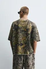 Sudadera relaxed fit con estampado de hojas en tonos verde, marrón y negro. Tiene cuello redondo, manga corta y bajo irregular con aberturas laterales.