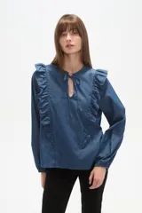 Blusa de tela tipo jean (denim) color azul medio, de corte holgado, con volados largos que recorren el frente y mangas largas con puños ajustados. Presenta un escote con abertura en V y lazos para anudar.