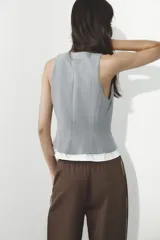 Chaleco gris con rayas verticales, escote en pico y cierre frontal con botones. Detalle de camisa blanca en el bajo.