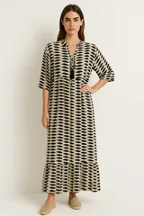 Vestido largo estampado, color beige y negro, con mangas tres cuartos y cuello mao con abertura en V anudada con borlas negras. Presenta talle ajustado con elástico y falda con volado en el ruedo.