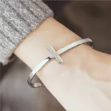 Pulsera rígida abierta de acero quirúrgico color plateado, con un dije de cruz en el centro.