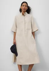 Vestido largo de lino color crema con finas rayas verticales. Tiene cuello mao, mangas cortas anchas, botonadura frontal oculta y un bolsillo en el pecho.